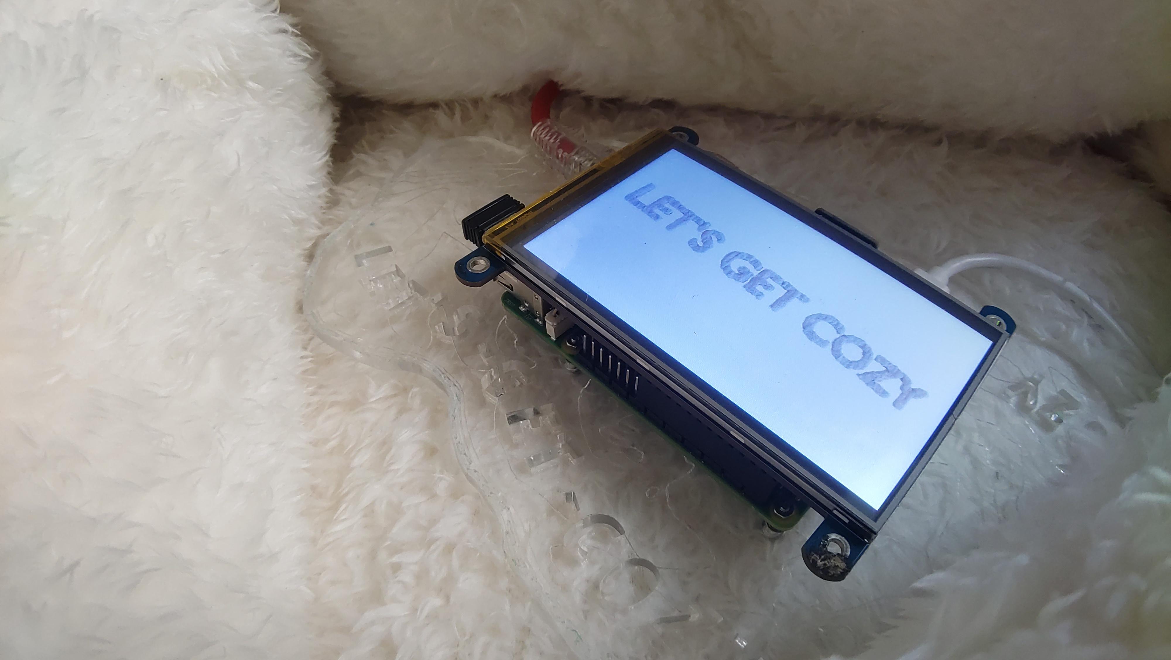 A close up of the Cozy-Cloud server sitting within the cloud pillows with its background sharing the words "Let's Get Cozy". The server sits of the tactile base with a textural cloud engraved on it and the words "let's get cozy" cut out of it..
