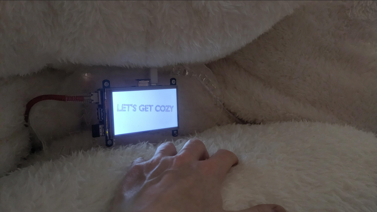 In among a cluster of Cozy-Cloud pillows, the small server's screen illuminates a hand reaching towards it. The screen they are reaching towards has written on it "Let's Get Cozy".